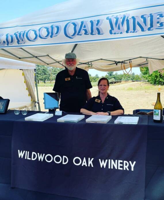 Anita and Tony Matlock staffing the Wildwood Oak Winery booth at an outdoor wine tasting event, with wine bottles displayed on the table.